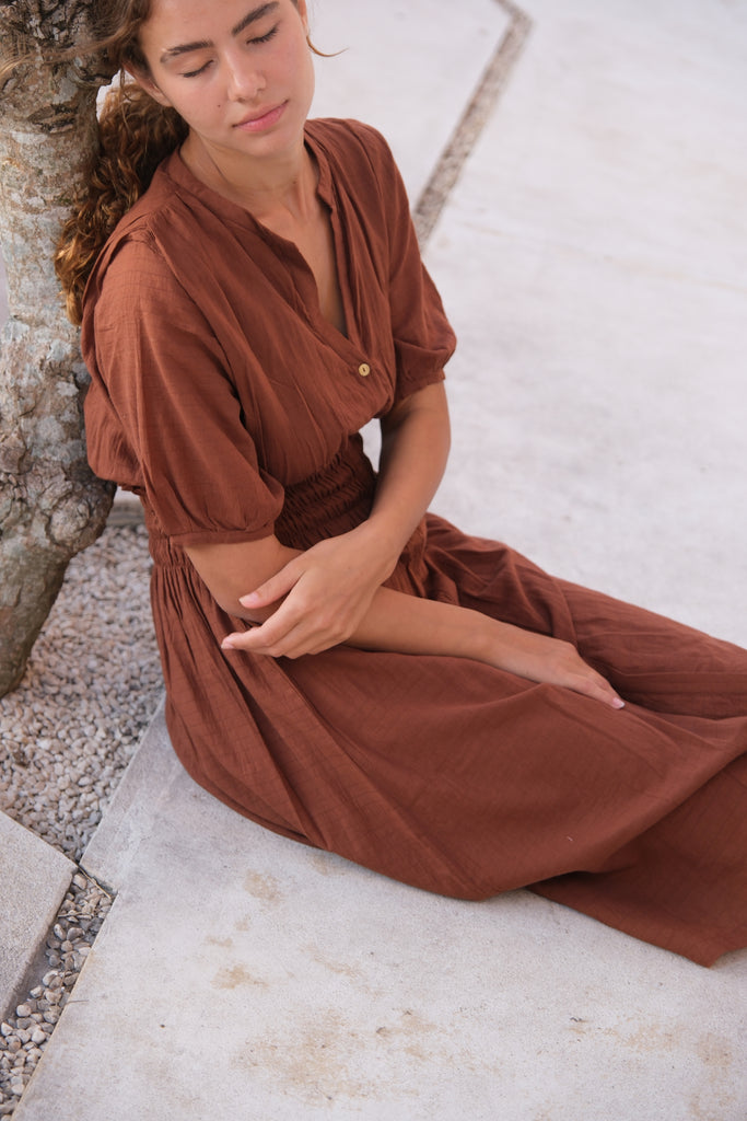 Introducing our Brown Handwoven Midi Dress, worn by a model standing beside a tree. This dress features intricate hand detailing and puff sleeves, perfect for adding a touch of elegance to your everyday look. Sustainably crafted from soft, breathable cotton, it combines style and comfort effortlessly.